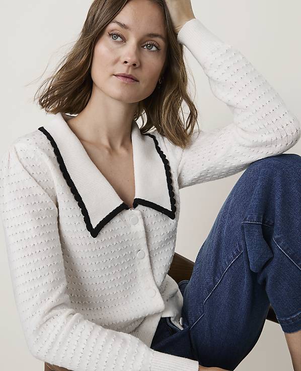A woman in a white collared cardigan and blue jeans sits against a blurred background.