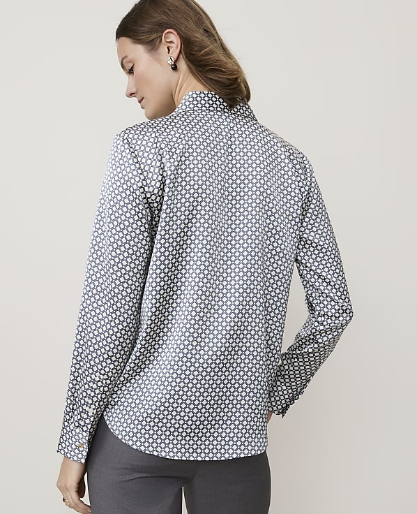 A woman in a patterned shirt stands against a plain, light-colored background.