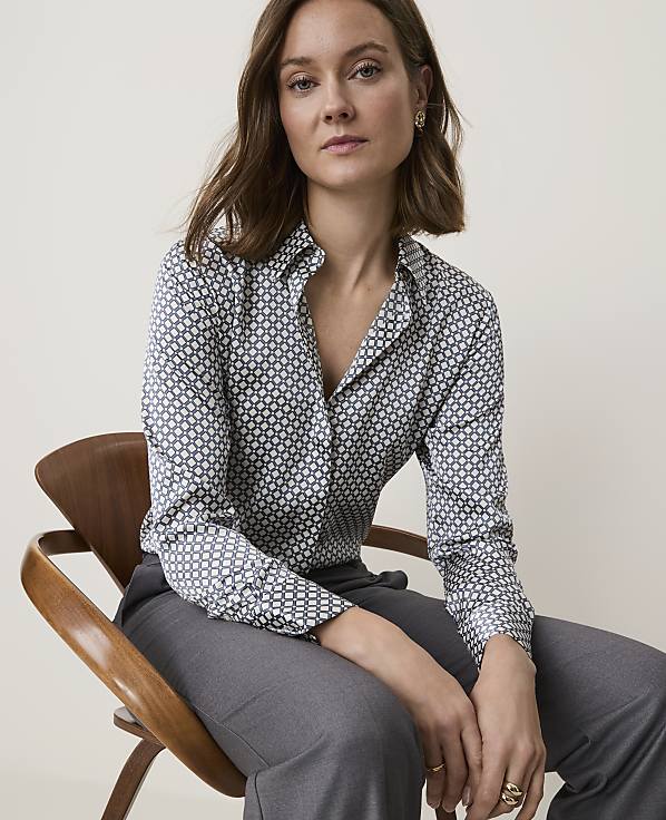 A woman in a patterned blouse and gray pants sits on a wooden chair against a plain, light background.
