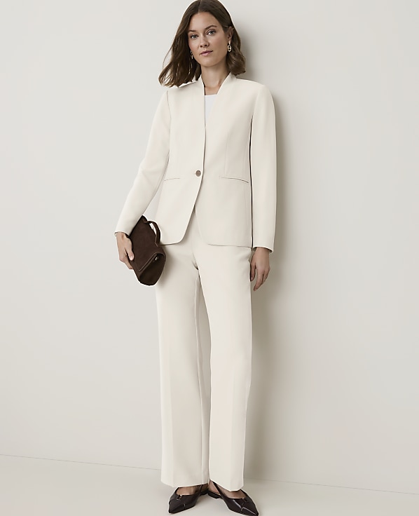 A woman in a cream suit and dark shoes holds a brown clutch in front of a plain white background.