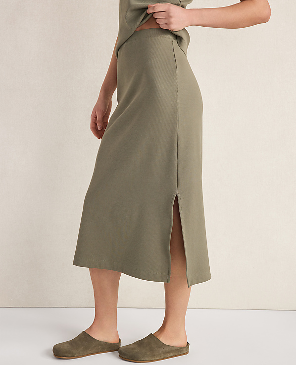 A person wearing a ribbed olive green midi skirt and clogs stands against a neutral, textured background.