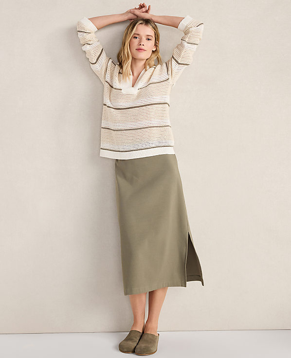 A woman in a striped sweater and olive skirt poses against a plain wall.