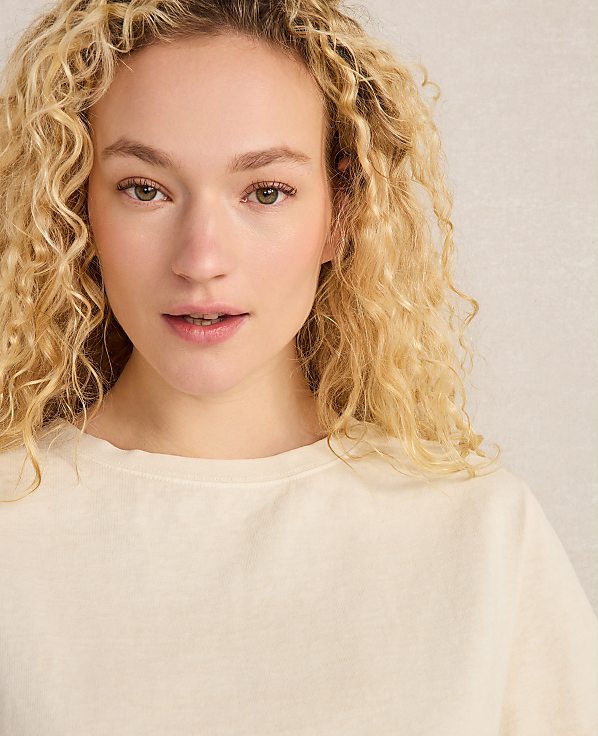 A young woman with curly blonde hair and fair skin looks forward, wearing a cream-colored shirt against a textured, light-colored background.