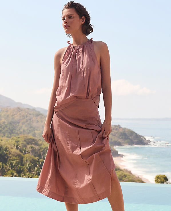A woman in a pink halter dress stands by a pool with a scenic ocean and mountain view in the background.
