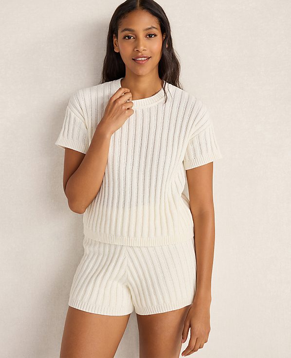 A woman in a cream-colored cable-knit top and shorts stands against a plain, textured cream background.