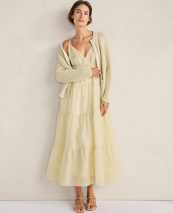 A woman in a light yellow tiered dress and a cream cardigan stands against a plain, light-colored wall.