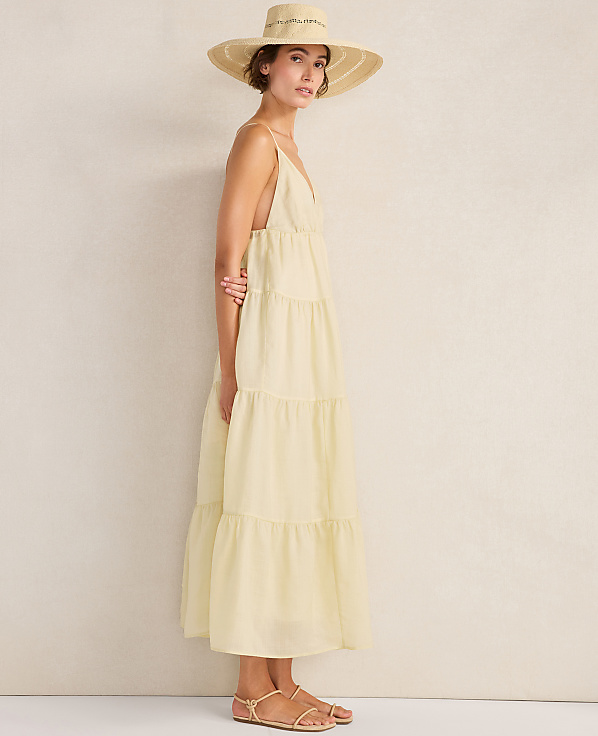 A woman in a light yellow tiered dress and a wide-brimmed hat stands against a plain, textured background.