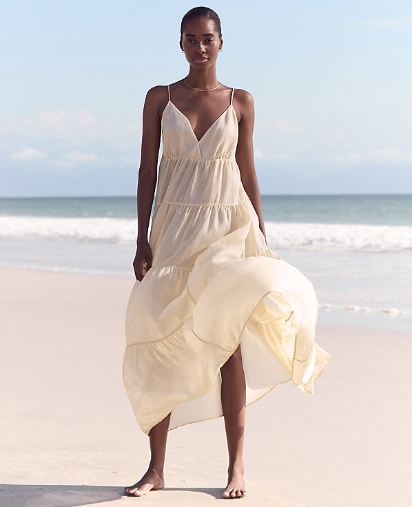 A woman in a flowing cream dress stands on a sandy beach with gentle waves and a clear sky in the background.