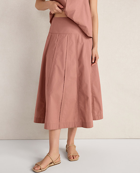 A person stands in a dusty rose pleated skirt and simple sandals against a textured off-white background.