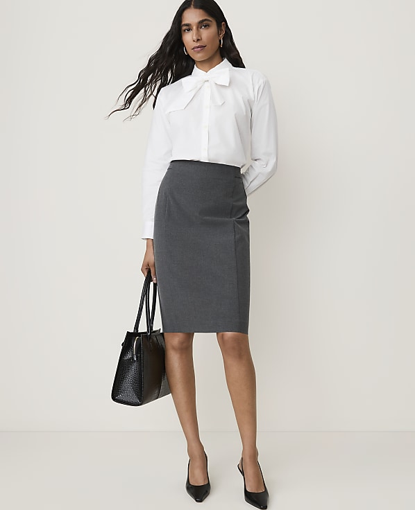 A woman in a white blouse and gray skirt stands against a plain white background, holding a black handbag.