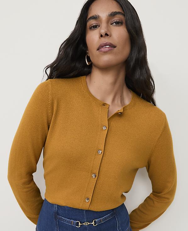 A woman in a mustard-colored cardigan stands against a plain, light background.