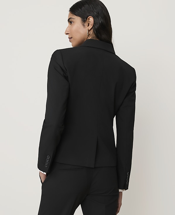 A woman in a black blazer and pants stands against a plain, light-colored background.