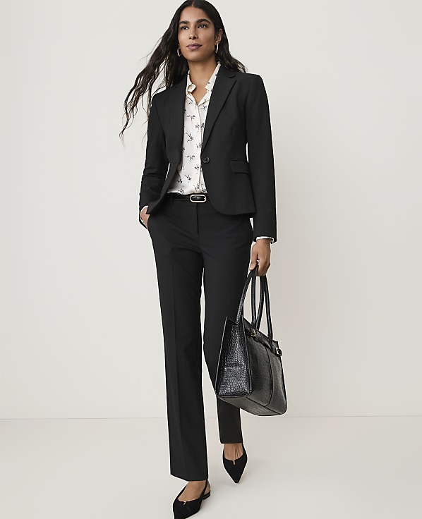 A woman in a black suit and patterned blouse stands with a black tote bag against a plain white background.