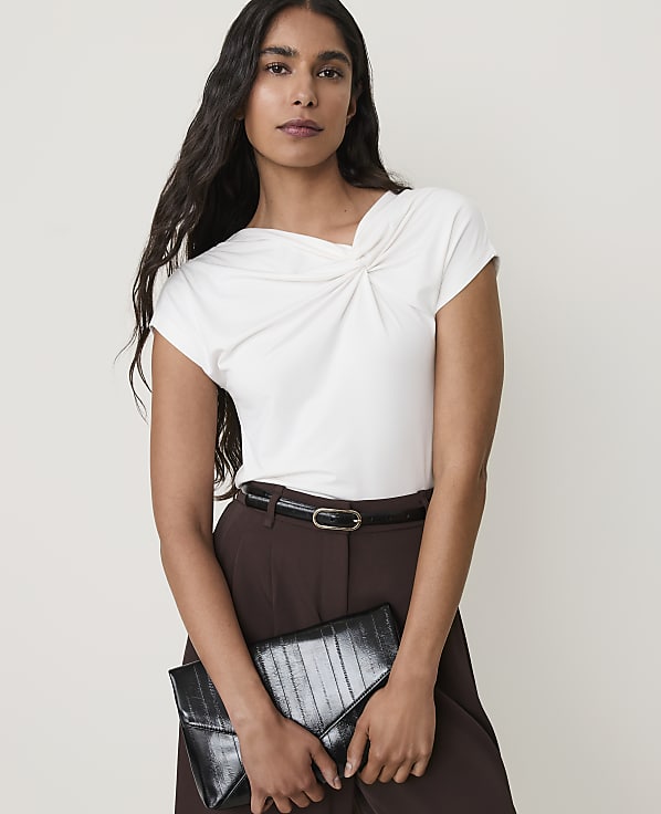 A woman in a white top and brown pants holds a black clutch in front of a plain background.