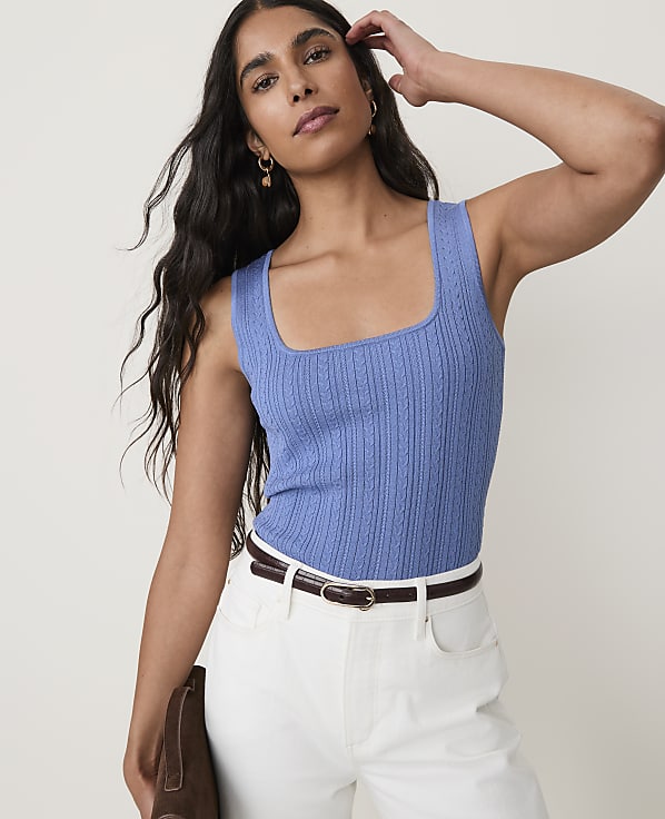 A woman in a blue knit top and white pants stands against a plain white background, holding a brown clutch.