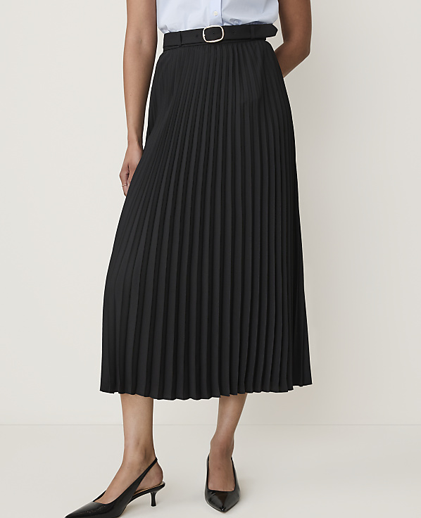 A person models a black pleated midi skirt with a belt and black slingback heels against a plain white background.