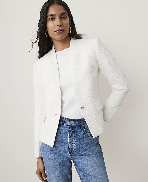 A woman in a white tweed blazer and blue jeans stands against a plain, light background.