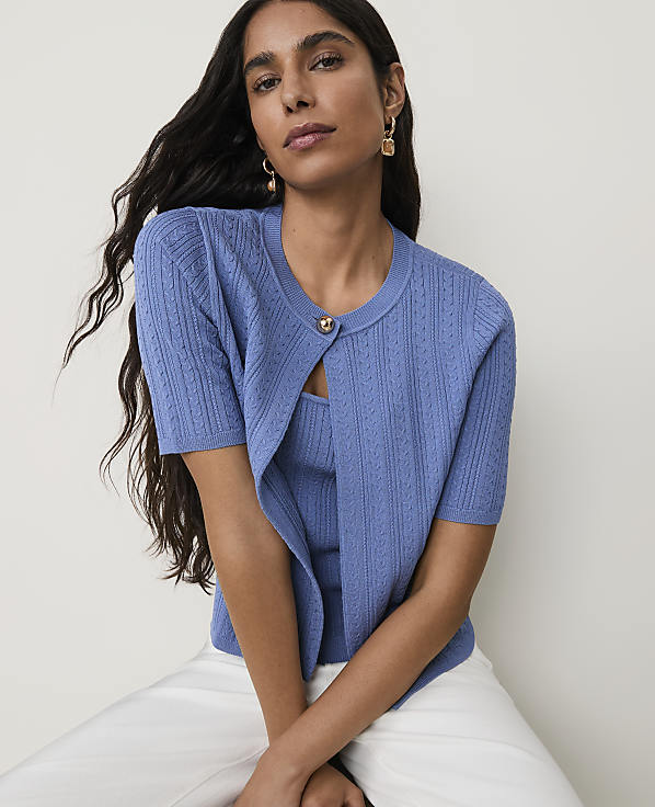 A woman with long dark hair wears a blue cardigan and tank top with white pants, against a plain white background.