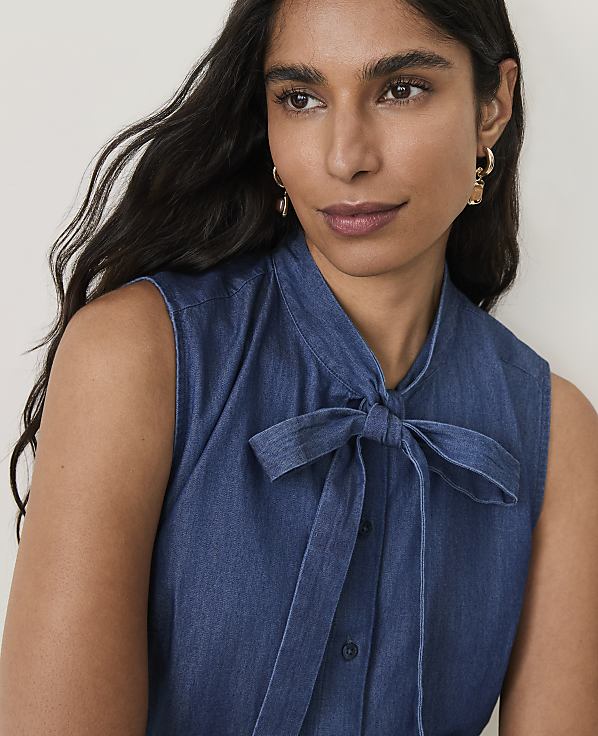 A woman with dark hair and gold earrings poses in a denim shirt with a bow tie in the foreground, against a plain, light background.