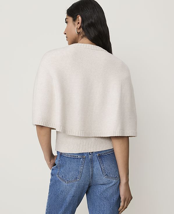 A woman in a cream-colored capelet and blue jeans stands against a plain white background.