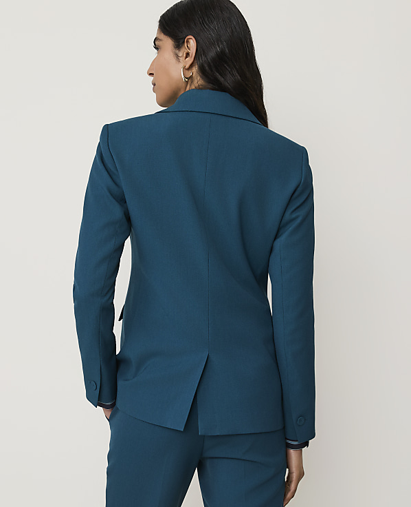 A woman in a teal blazer and pants stands with her back to the camera against a plain white background.
