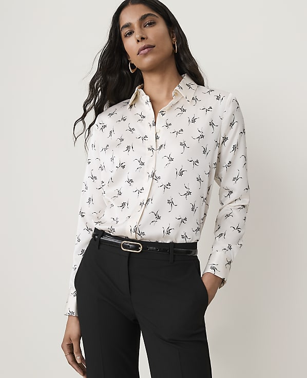 A woman in a patterned blouse and black pants stands against a plain background.
