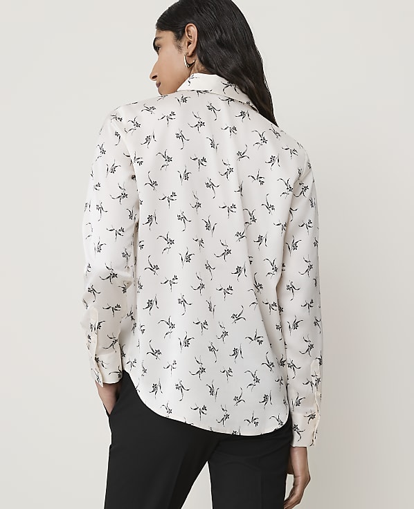 A woman in a white patterned shirt and black pants stands against a plain, light-colored background.