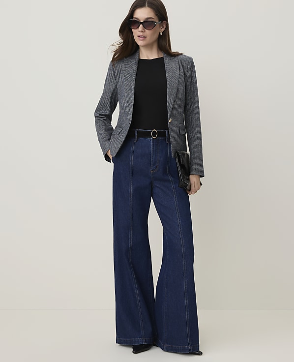A woman in a gray blazer and wide-leg jeans stands against a plain, light-colored background.
