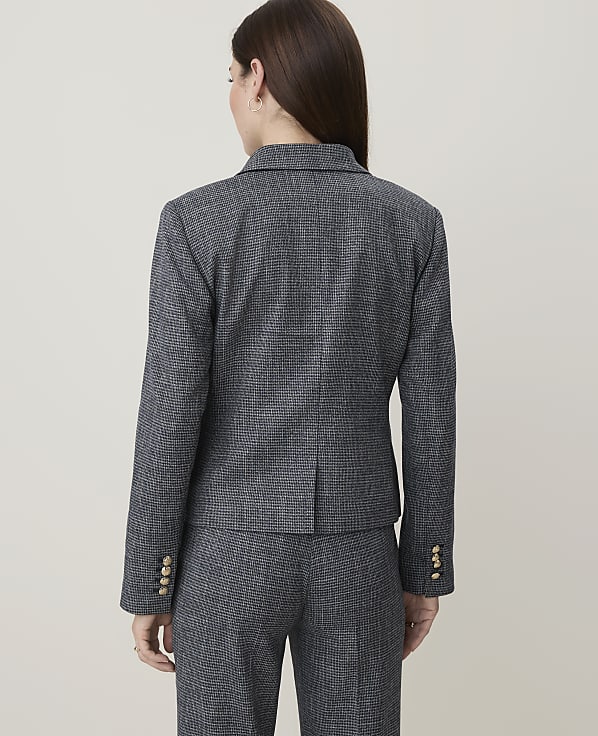 A woman in a grey patterned blazer and pants stands against a plain grey background.