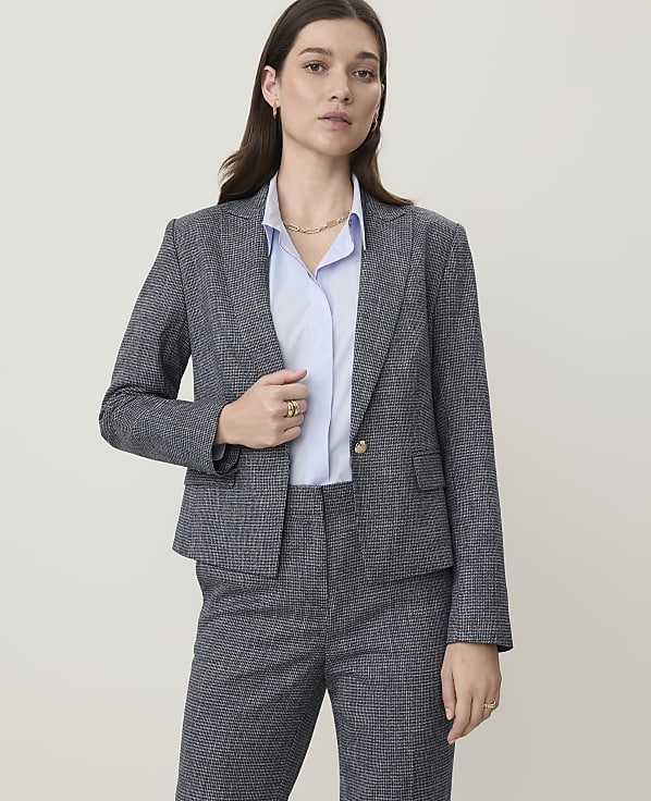 A woman in a grey patterned suit stands against a plain, light-colored background.