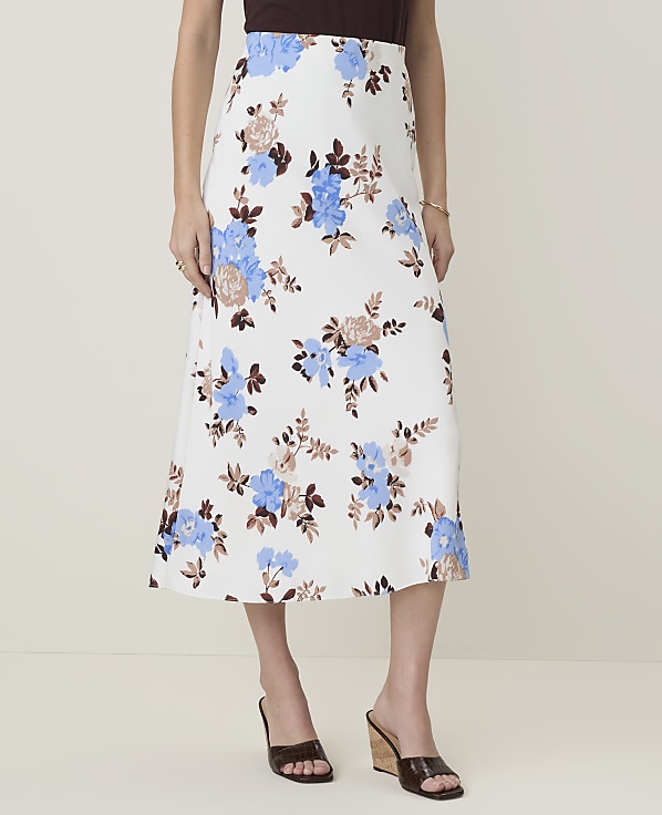 A woman models a white floral midi skirt with blue and brown flowers, paired with brown wedge sandals, against a plain, light-colored background.