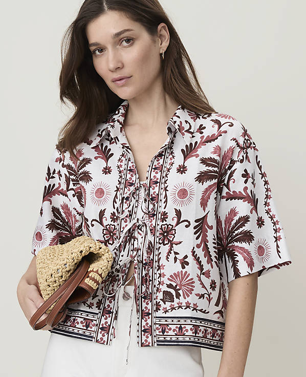 A woman in a patterned shirt holds a straw bag in front of a plain background.
