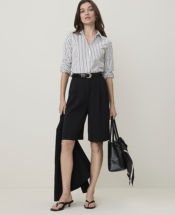 A woman in a striped shirt and black culottes stands against a plain, light background, holding a black blazer and a tote bag.