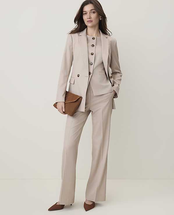 A woman in a beige pantsuit and vest holds a brown clutch in front of a plain, light-colored background.