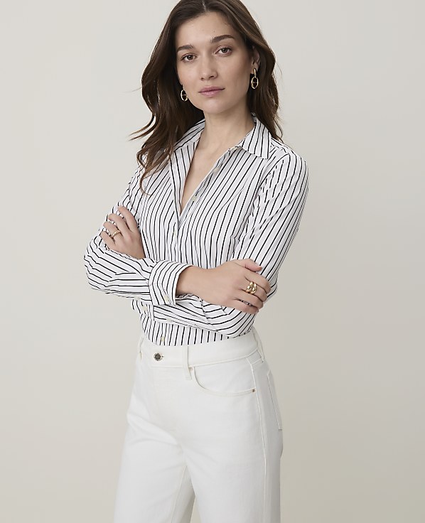 A woman in a striped shirt and white pants stands with her arms crossed against a plain, light background.