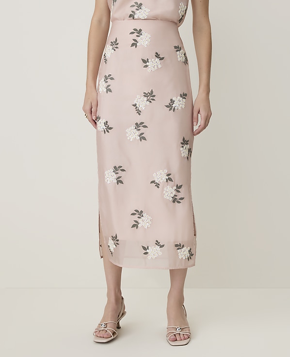 A person stands in front of a plain background, wearing a light pink floral embroidered midi skirt and light-colored sandals.