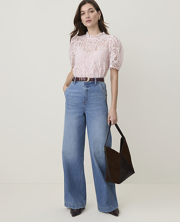 A woman in a pink lace top and wide-leg jeans stands against a plain, light background, holding a dark brown handbag.