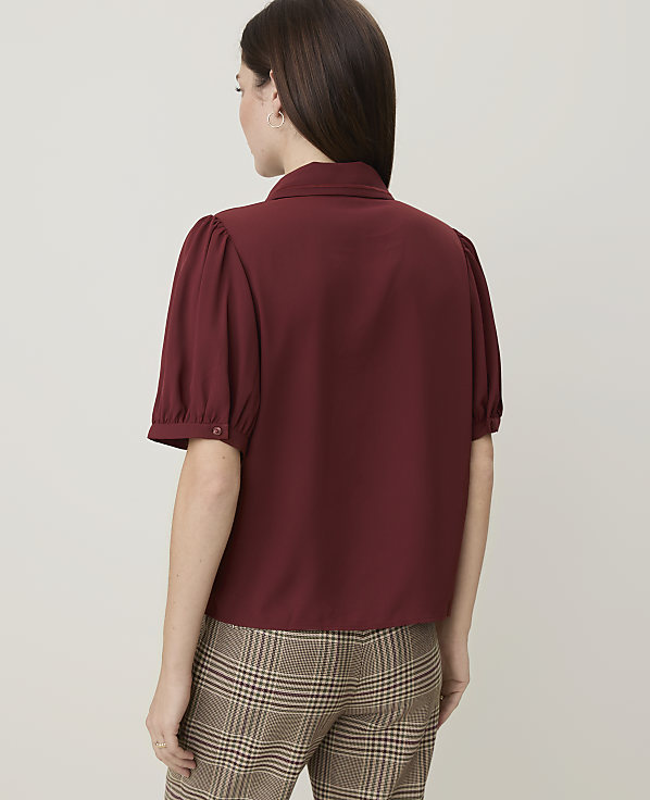 A woman in a maroon puff-sleeve blouse and plaid pants stands against a plain background.