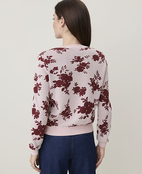 A woman in a pink floral sweater and blue jeans stands against a plain, light background.