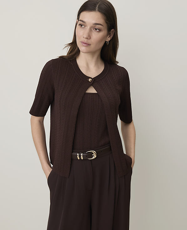 A woman in a brown cardigan and matching top stands against a plain, light-colored background.