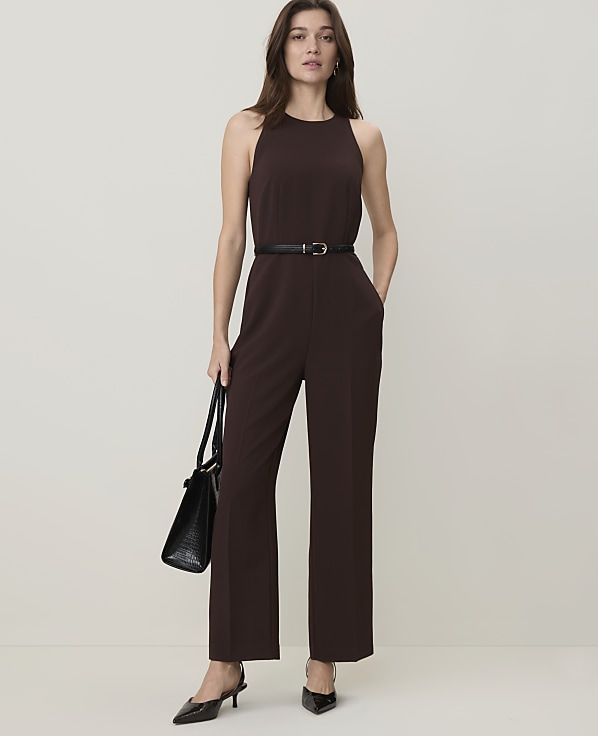 A woman in a brown jumpsuit and black heels stands with a black handbag against a plain, light brown background.