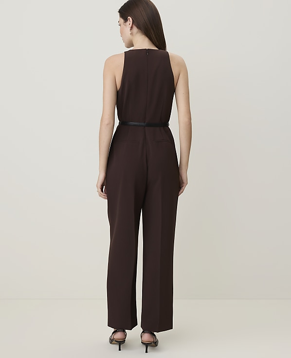 A woman in a brown jumpsuit with a black belt stands against a plain, light-colored background.
