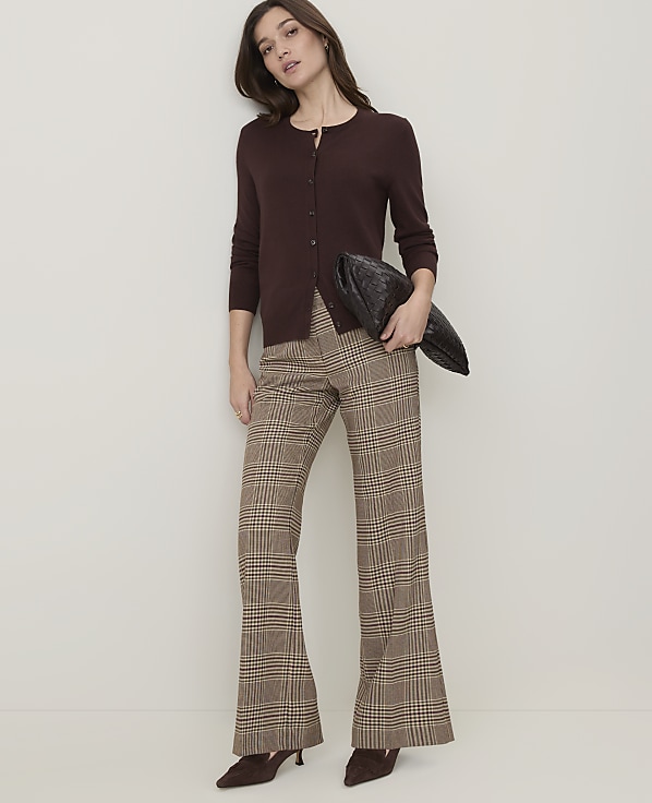 A woman in a brown cardigan and plaid flared pants stands against a plain, light-colored wall, holding a dark clutch.