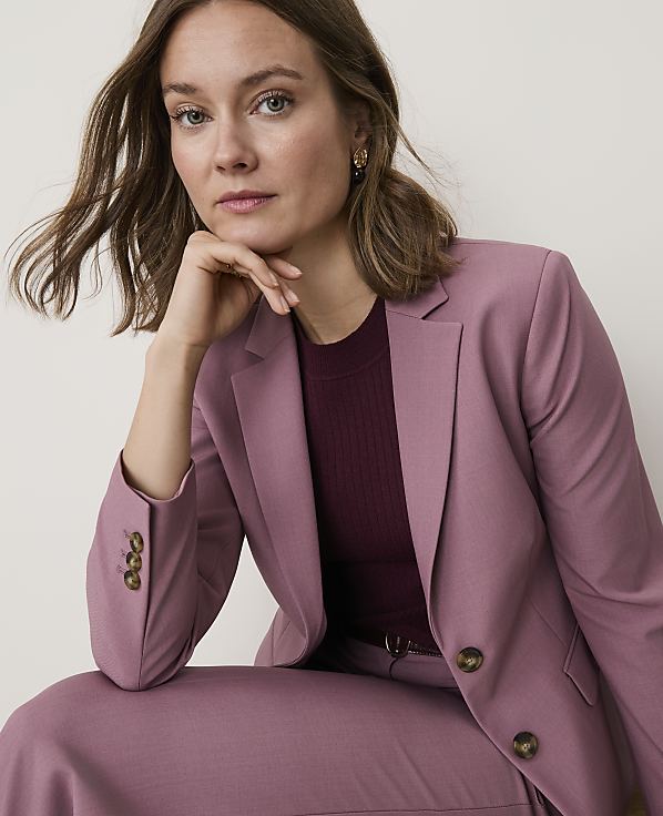 A woman in a mauve blazer and matching pants rests her chin on her hand against a plain, light background.