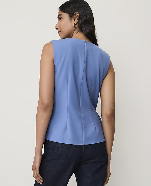 A woman in a blue sleeveless top and dark pants stands against a plain white background.