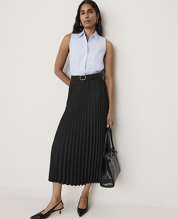 A woman in a light blue sleeveless shirt and a black pleated skirt stands against a plain white background, holding a black handbag.
