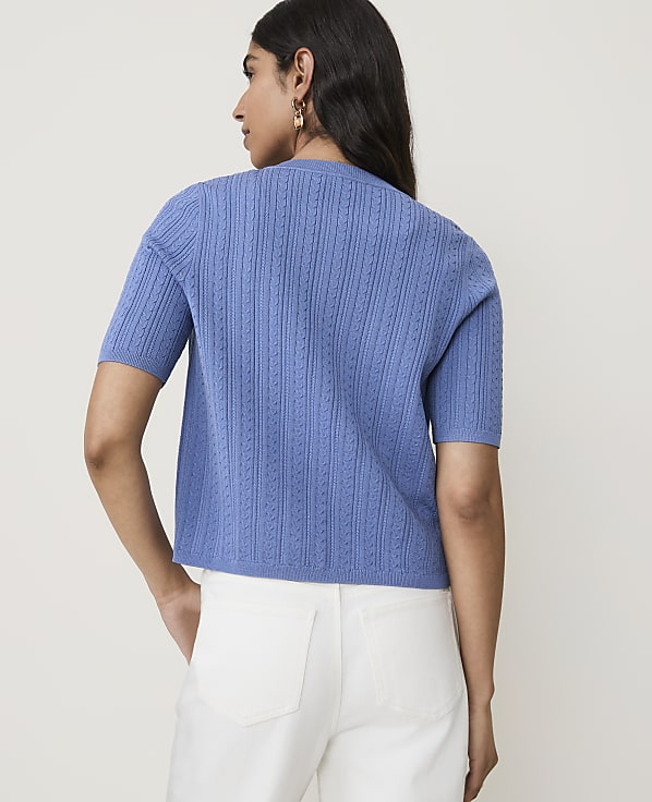 A woman in a blue cable-knit sweater and white pants stands against a plain white background.