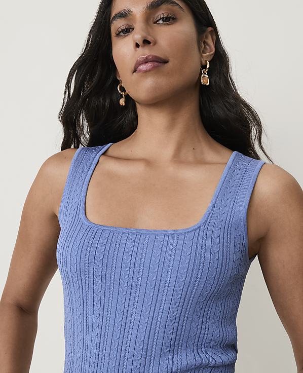 A woman with dark hair and earrings wears a blue cable-knit tank top against a plain background.