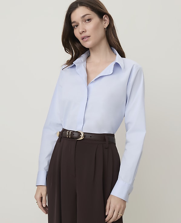 A woman in a light blue collared shirt and dark brown pants stands against a plain, light gray background.