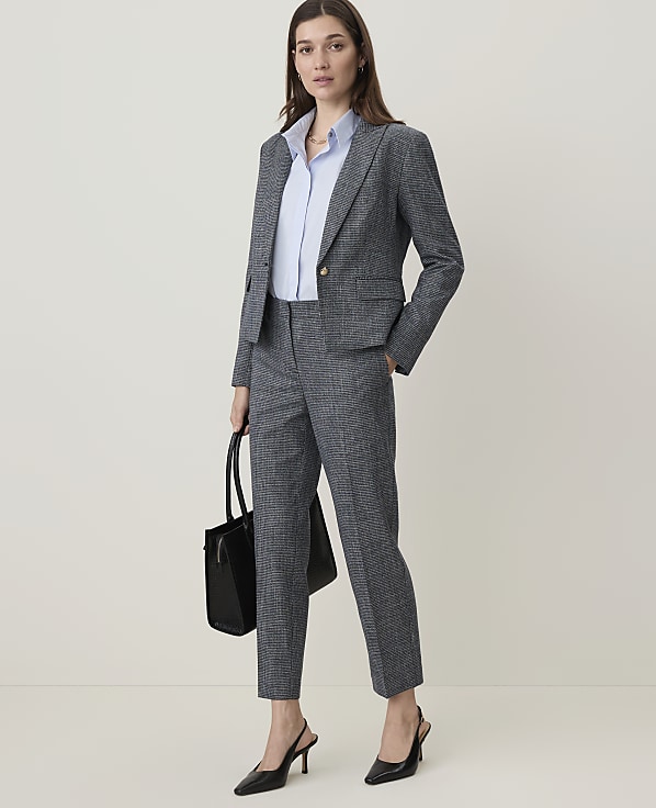 A woman in a grey suit and light blue shirt stands holding a black bag, with a plain, light grey background.